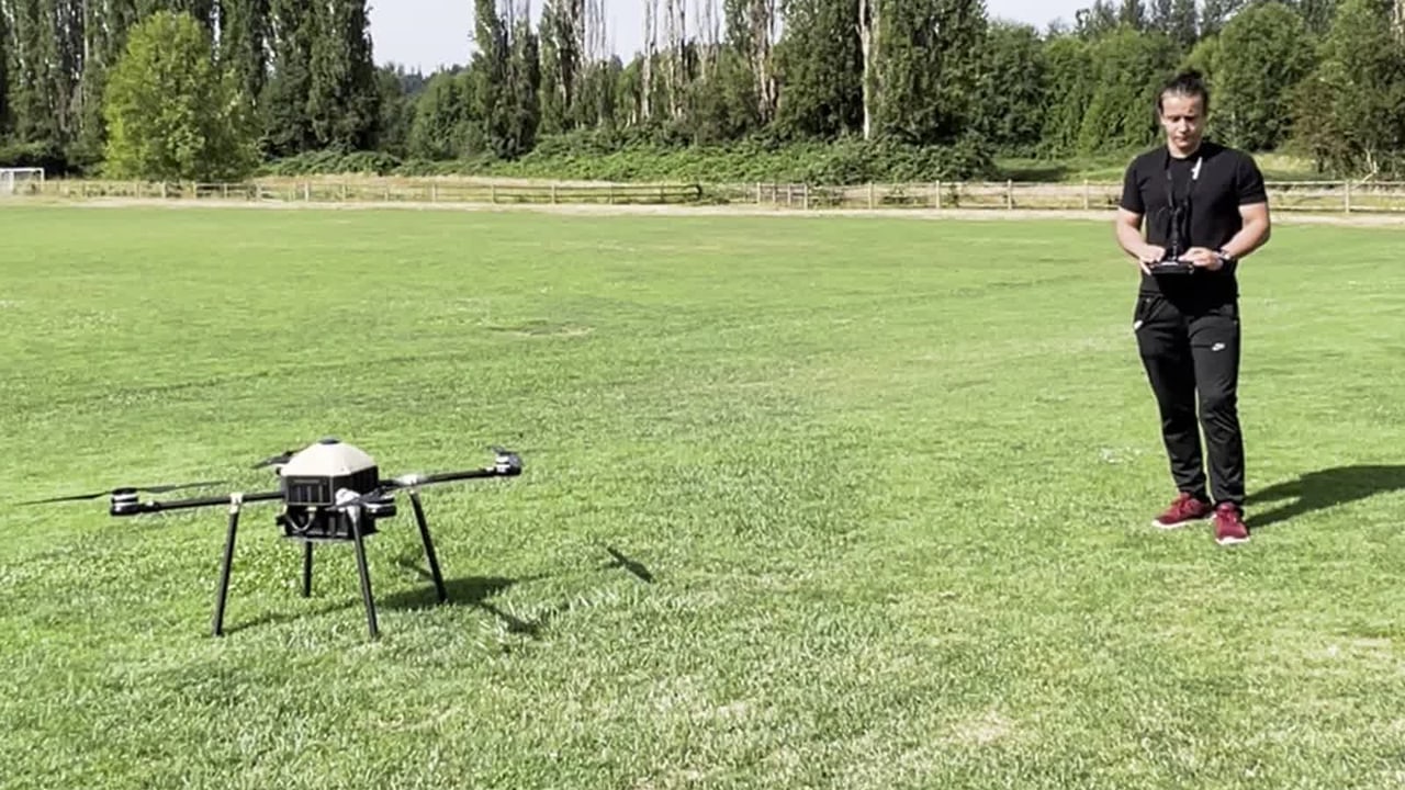 The aircraft airborne over the field with the safety pilot standing below, transmitter in hand, ready to take control